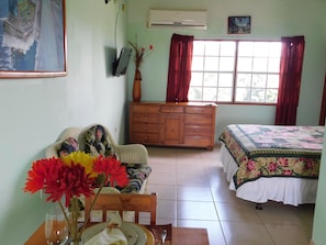 A room with a bed, a wooden dresser, a chair, a table with a vase of flowers, and a window with curtains.