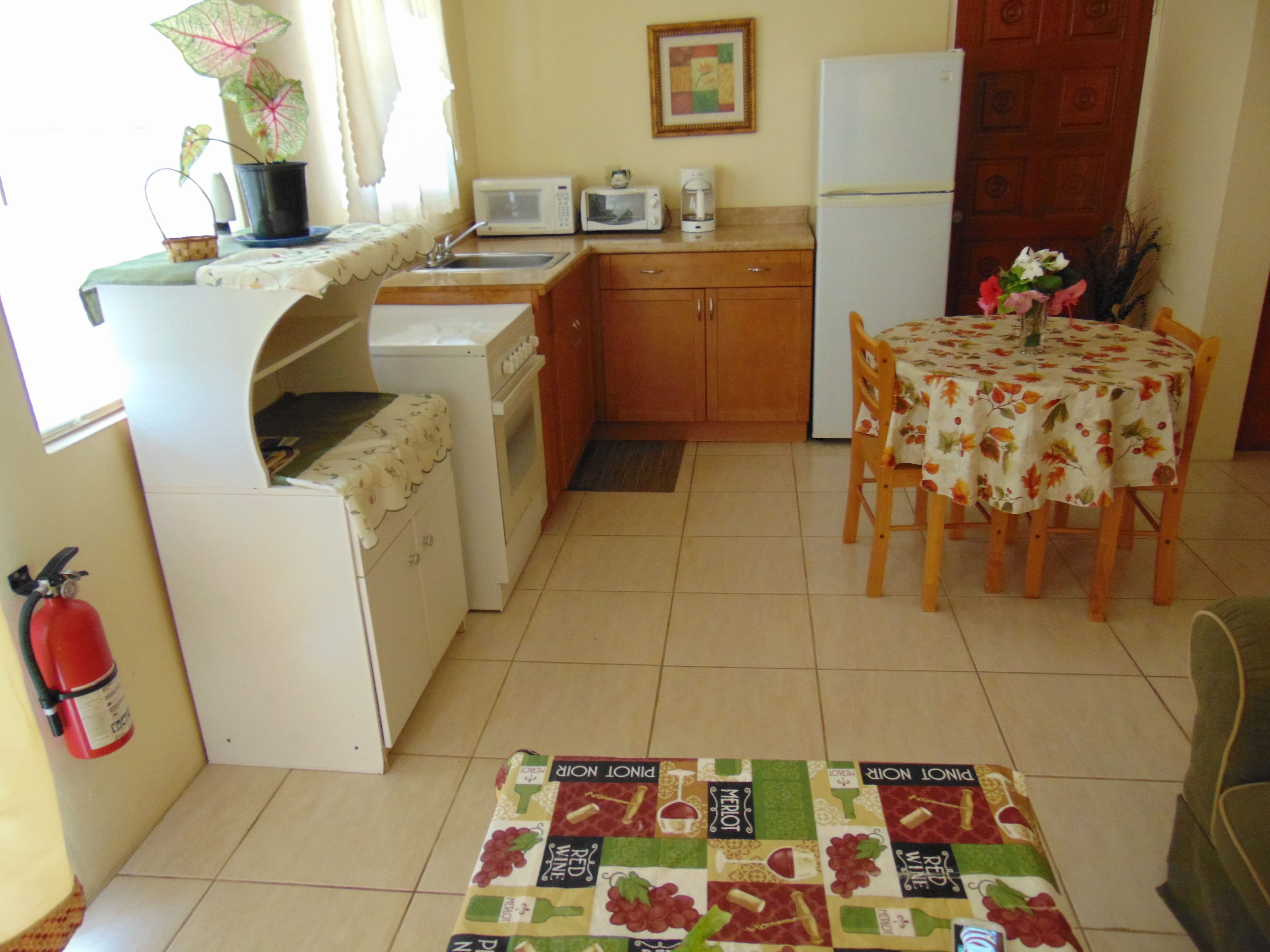 A compact kitchen with a microwave, toaster, and refrigerator. There's a dining table with a floral tablecloth and chairs. A small potted plant is on the counter.