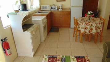 A compact kitchen with a microwave, toaster, and refrigerator. There's a dining table with a floral tablecloth and chairs. A small potted plant is on the counter.