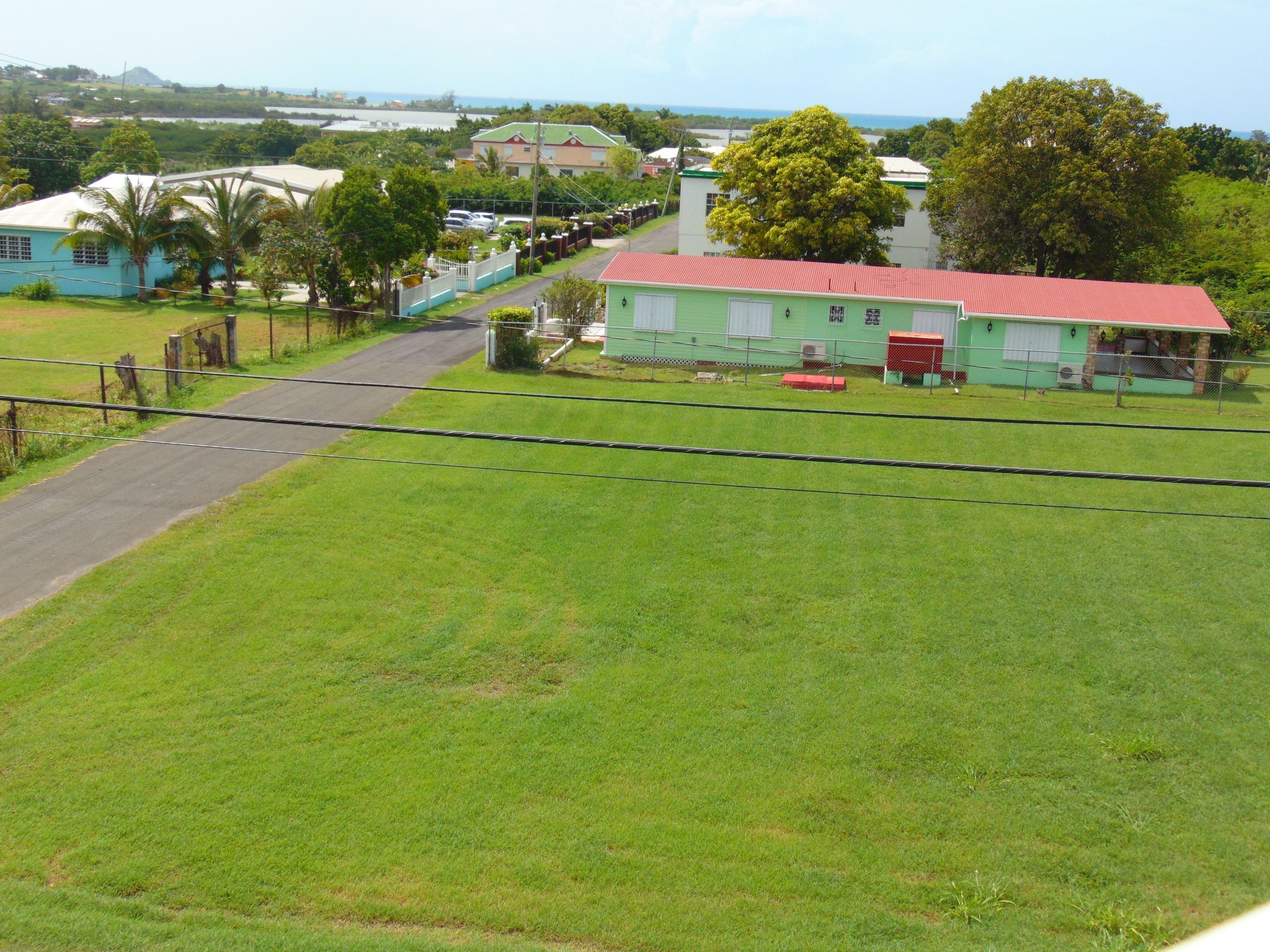 Um edifício verde com telhado vermelho, cercado por um quintal murado e uma estrada que leva até ele.