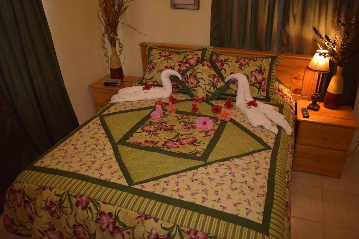 A bed with floral bedding, a nightstand with a lamp, and a vase with dried plants.