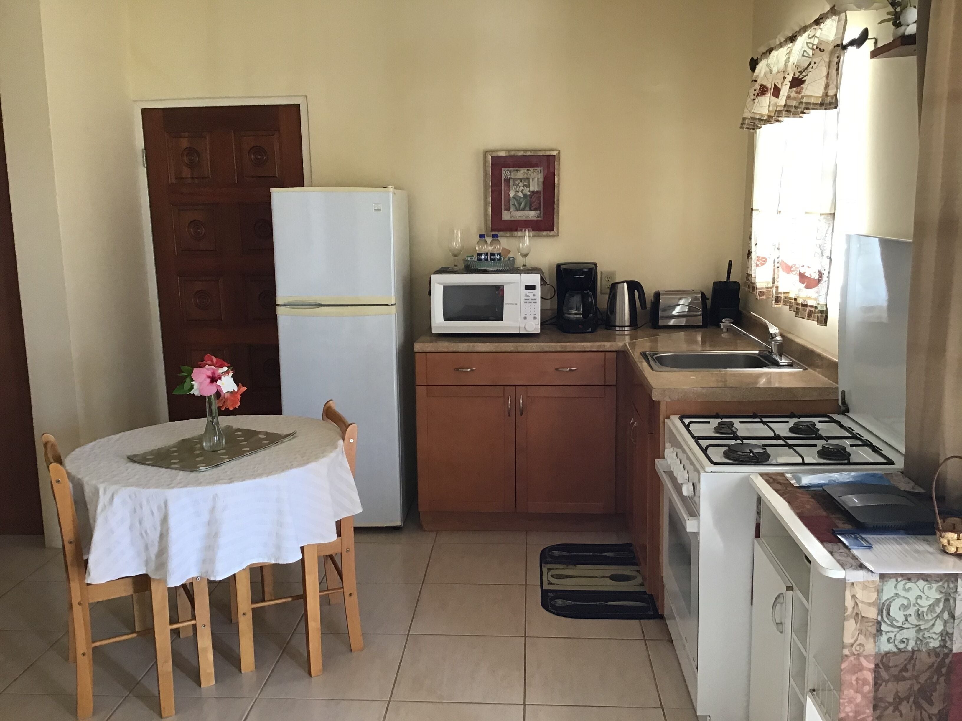 A compact kitchen with a refrigerator, microwave, toaster, and gas stove. There's a dining table with a chair and a vase of flowers.