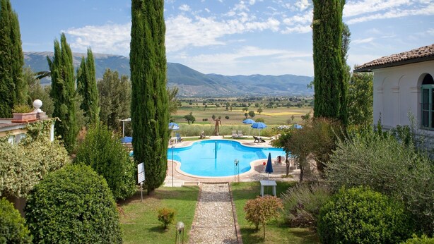 A swimming pool surrounded by tall cypress trees, with a view of distant mountains and a white building with a tiled roof. - Subretia Residenze di Campagna (Montefalco)