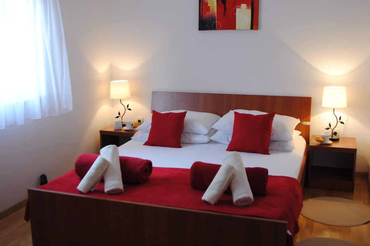 A neatly made bed with red and white bedding, two bedside tables with lamps, a painting on the wall, and a wooden floor.