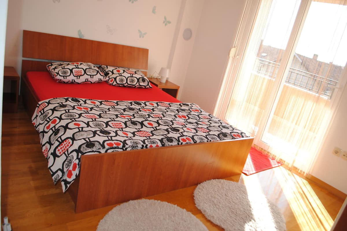 A bedroom with a wooden bed, red bedding, a nightstand, and a window with curtains.