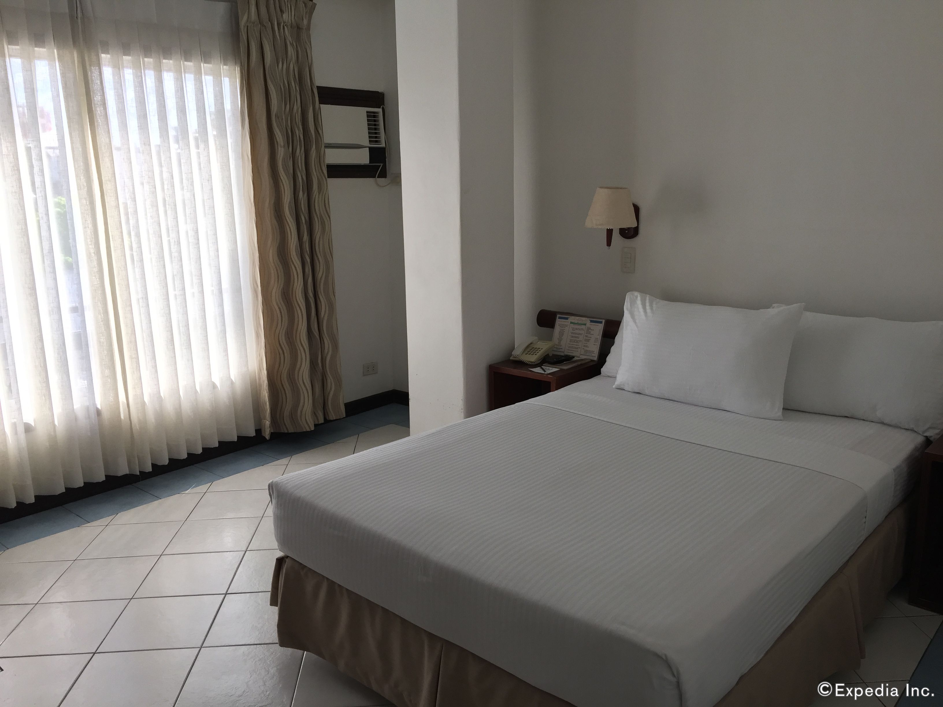 Una habitación de hotel con una cama grande, una mesita de noche con lámpara, un teléfono y un aire acondicionado de pared.