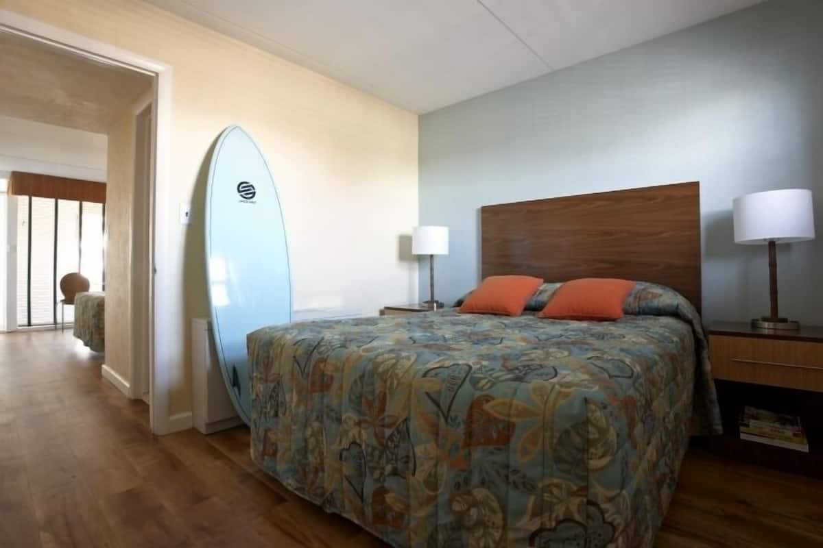 A bedroom with a bed, a nightstand, a lamp, and a surfboard leaning against the wall.