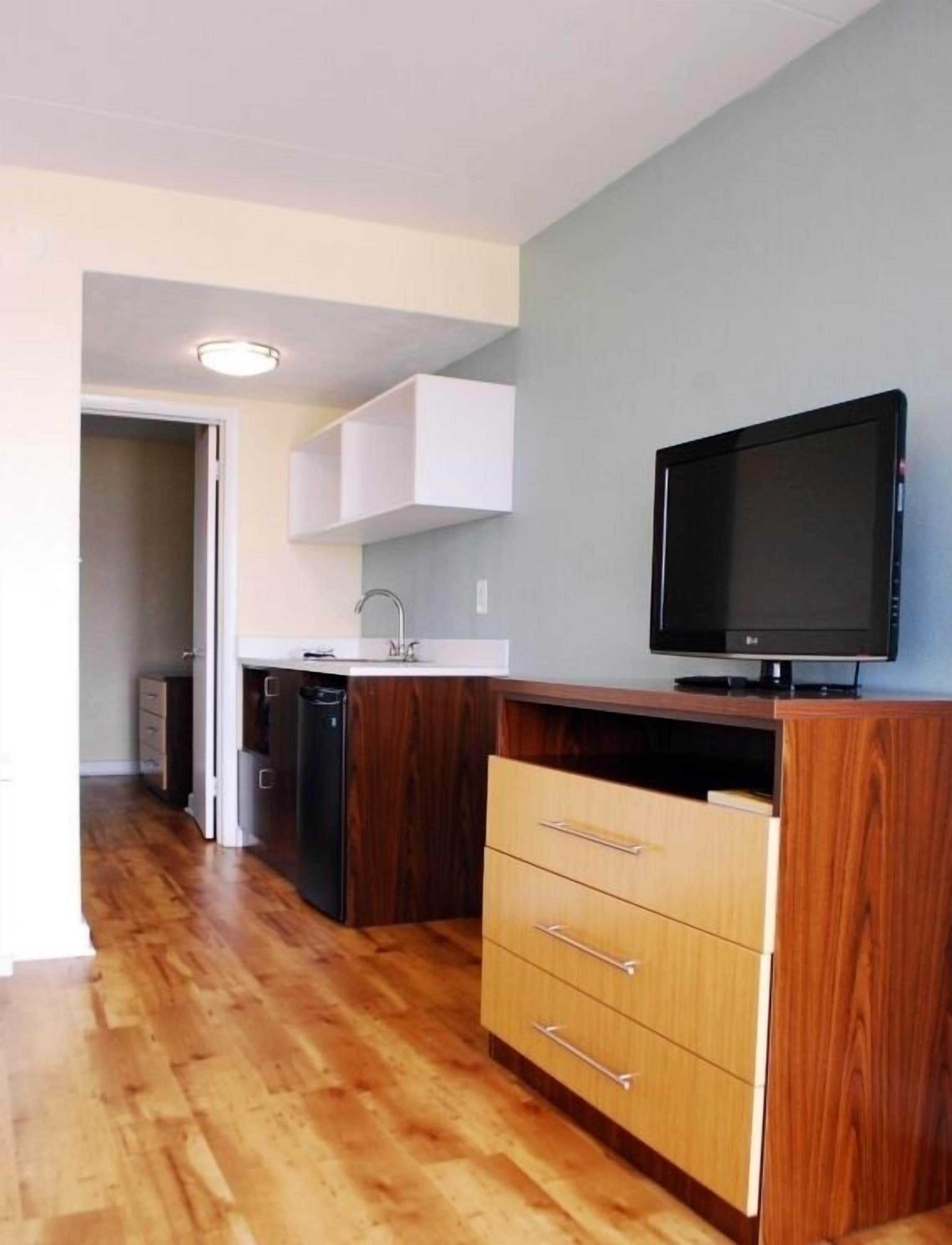 A compact kitchen with wooden cabinets, a built-in dishwasher, and a flat-screen TV on a wooden stand.