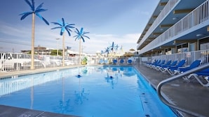 Una piscina con palmeras azules, sillas de descanso y un edificio con balcones.