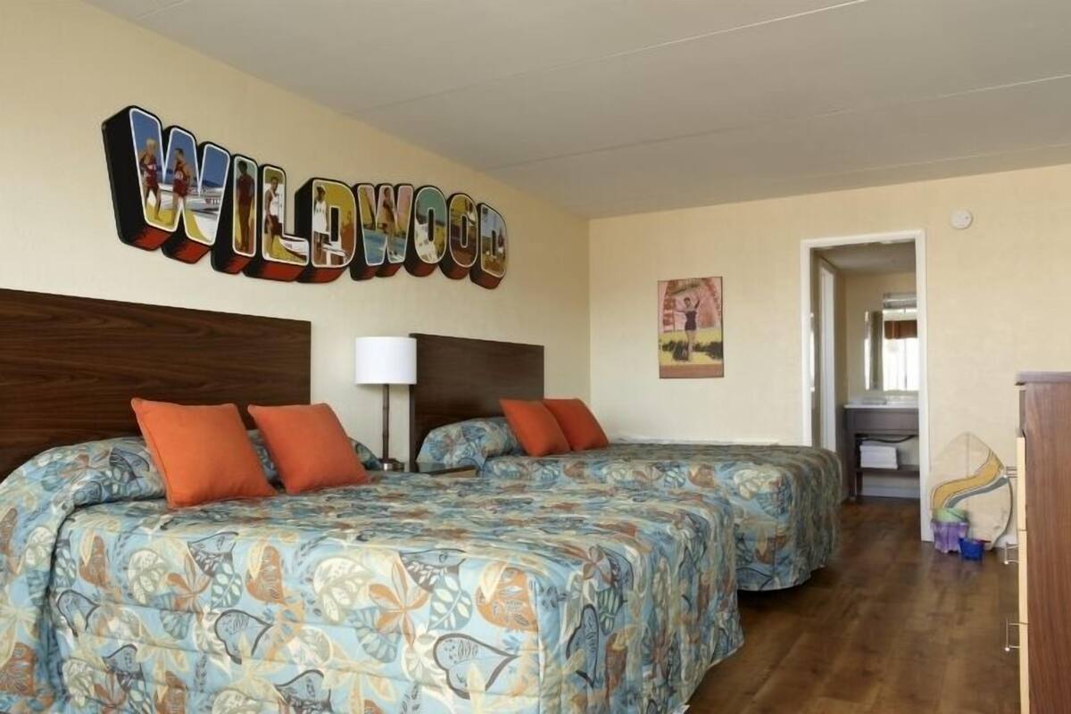 A hotel room with two beds, a wooden headboard, a wall sign reading "WILDWOOD", and a painting on the wall.