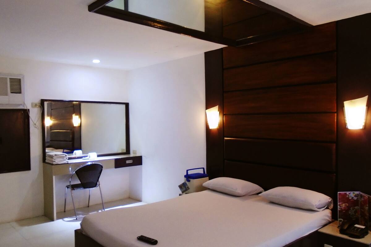 Quarto de hotel com cama, escrivaninha com cadeira, espelho e luminárias na parede.