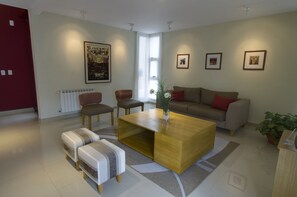 A modern living room with a sofa, coffee table, and ottomans. There is a radiator, a window, and framed pictures on the wall. - Quinta & Suites Apart Hotel (Mendoza)