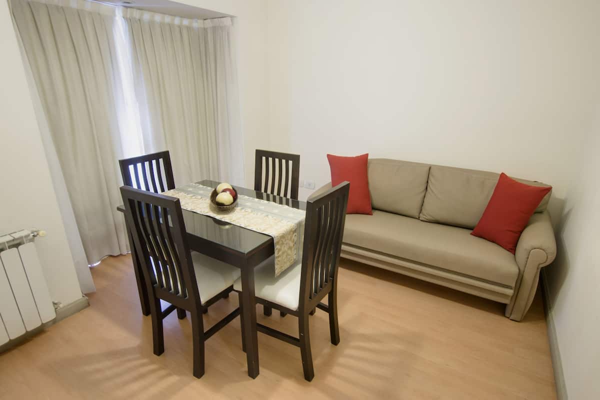 A dining area with a table and four chairs, a sofa with red cushions, and a radiator.