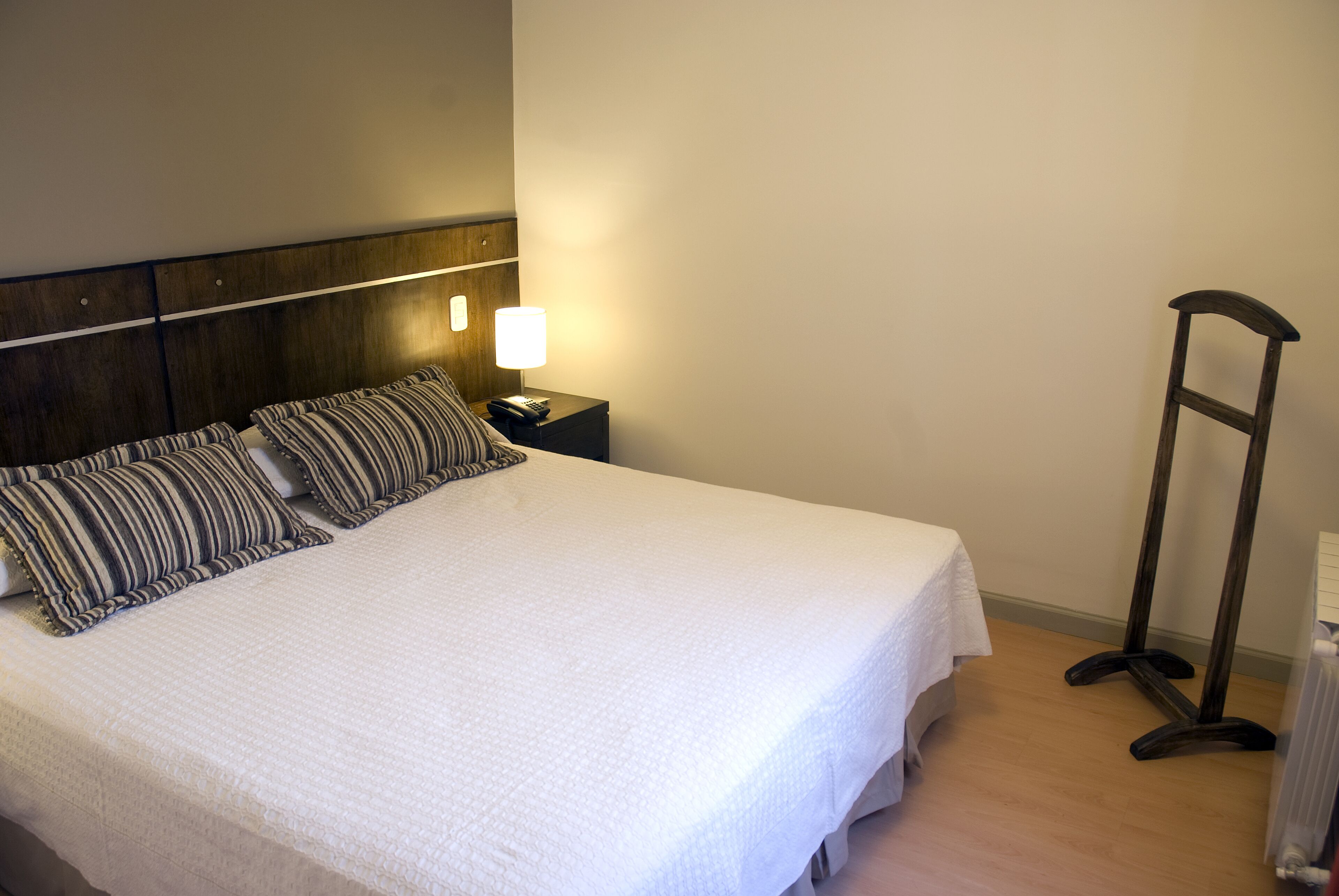 Una cama bien hecha con edredón blanco y almohadas a rayas, una lámpara de noche y una mesita de madera.