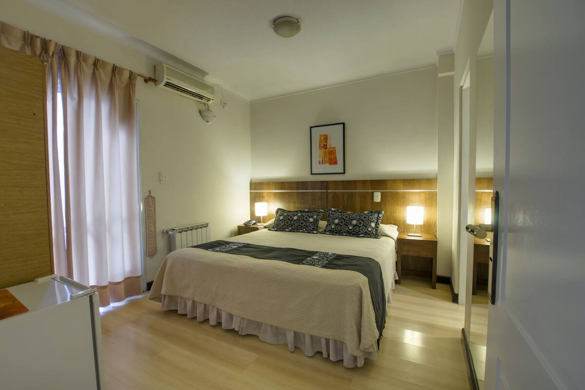A hotel room with a bed, bedside lamps, a headboard, a radiator, and a window with curtains.