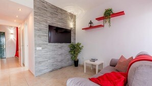 Un salon moderne avec un canapé gris, des coussins rouges, un téléviseur à écran plat fixé sur un mur texturé, une petite table d’appoint blanche et des étagères rouges murales ornées de plantes.