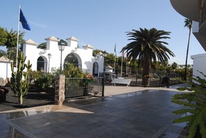 A courtyard with a white building, palm trees, and a flag. - Lovely Bungalows (San Bartolomé de Tirajana)