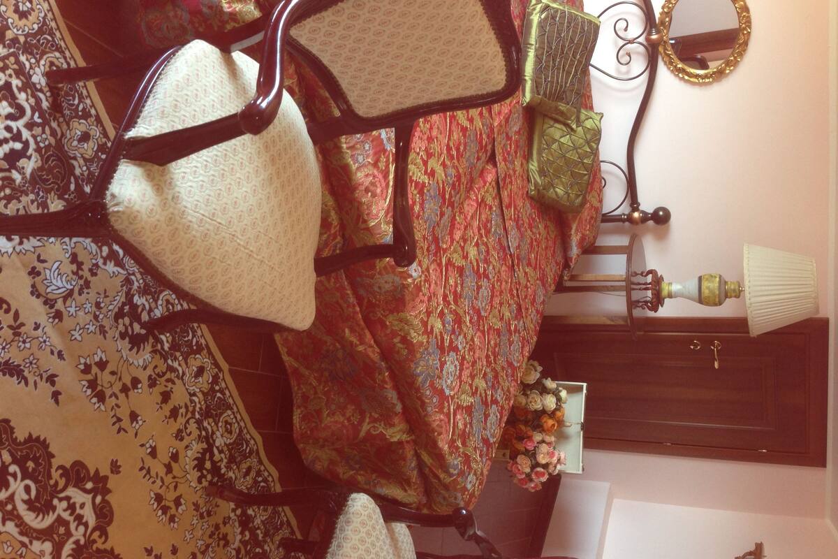 A room with a patterned carpet, a floral bedspread, a mirror, a lamp, and a small vase with flowers.