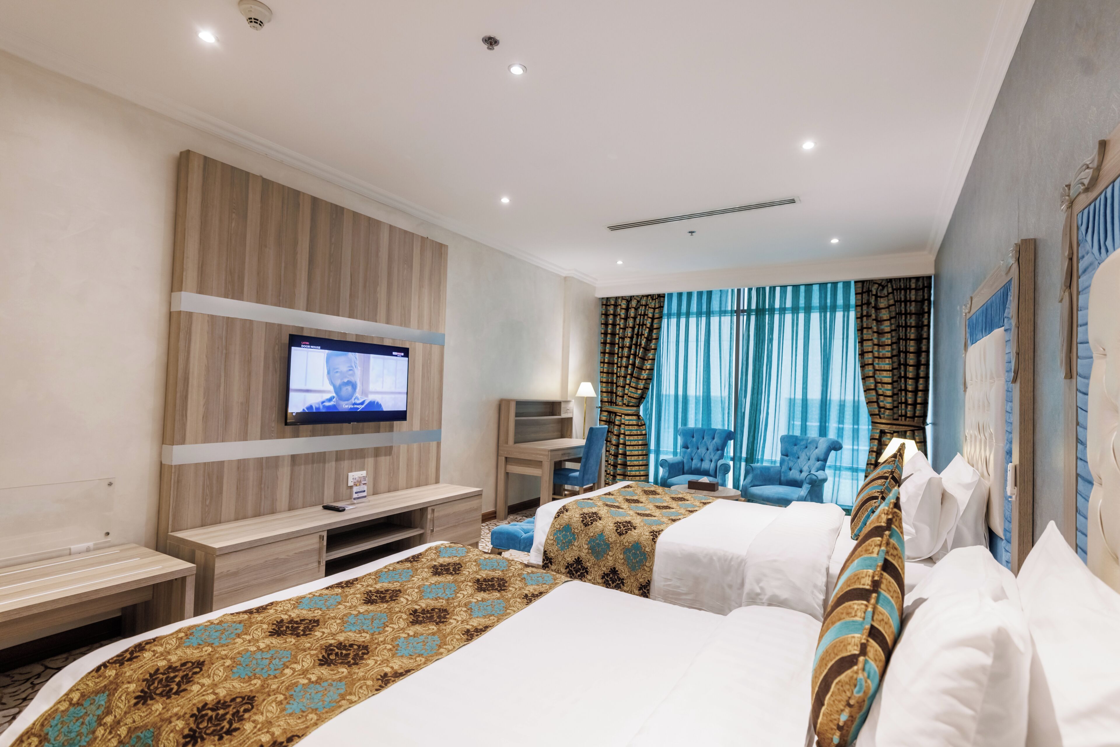 Habitación de hotel con dos camas, un escritorio, un televisor y vista al mar a través de amplias ventanas.
