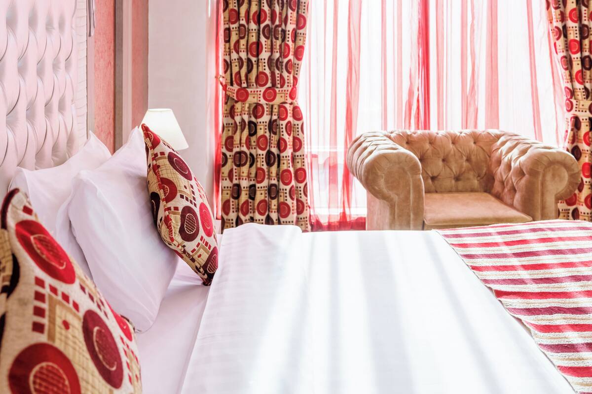 A hotel room with a large bed, a tufted headboard, a chaise lounge, and a window with sheer curtains.