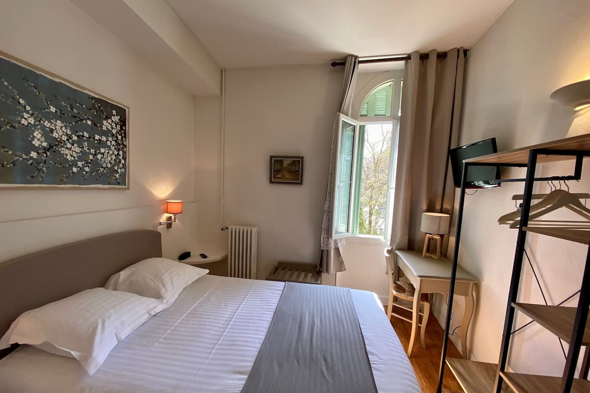 A hotel room with a bed, a desk, a chair, a radiator, a TV, and a painting on the wall.