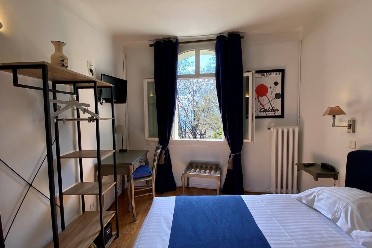 A bedroom with a bed, a desk, a chair, a radiator, a window with curtains, and a wall-mounted shelf with a vase.