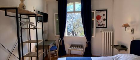 A bedroom with a bed, a desk, a chair, a radiator, a window with curtains, and a wall-mounted shelf with a vase.