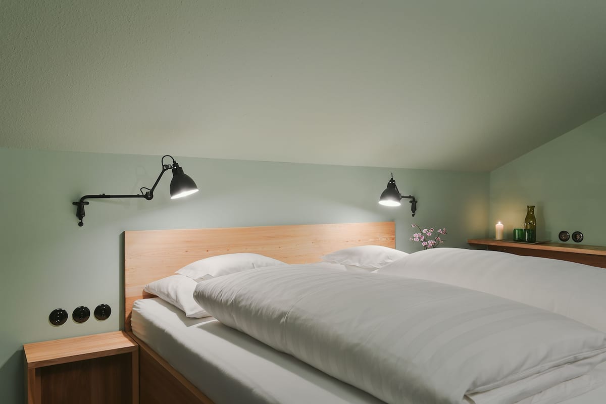 Una cama bien hecha con sábanas blancas en una habitación con paredes verdes, dos lámparas de noche y una mesita con un pequeño jarrón y una vela.