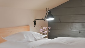 Ein ordentlich bezogenes Bett mit weißen Laken, eine Wandlampe und ein kleiner Blumenstrauß mit rosa Blumen.