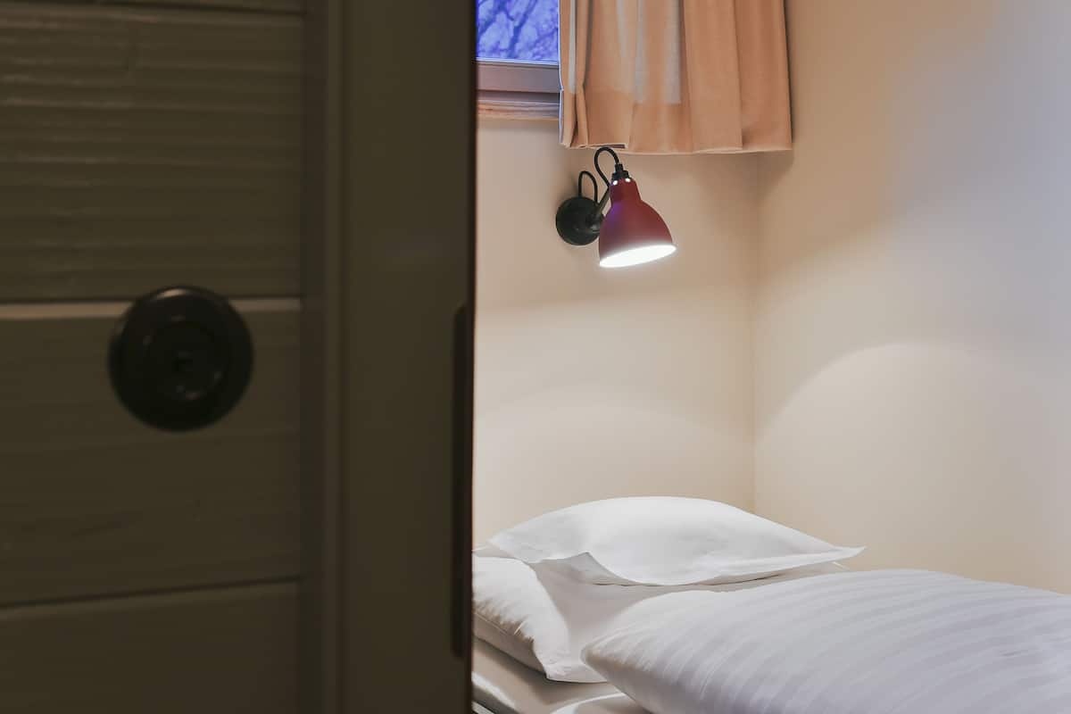 Un dormitorio con una cama individual, ropa de cama blanca, una lámpara de pared y una ventana con cortinas.