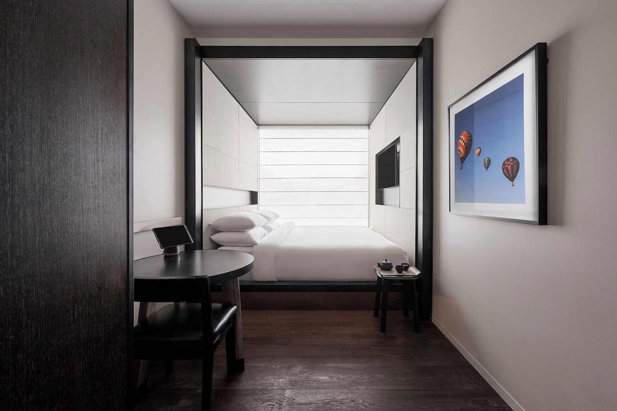 Une chambre d’hôtel compacte comprenant un lit, un bureau, une chaise, une petite table avec une théière et un tableau encadré au mur.