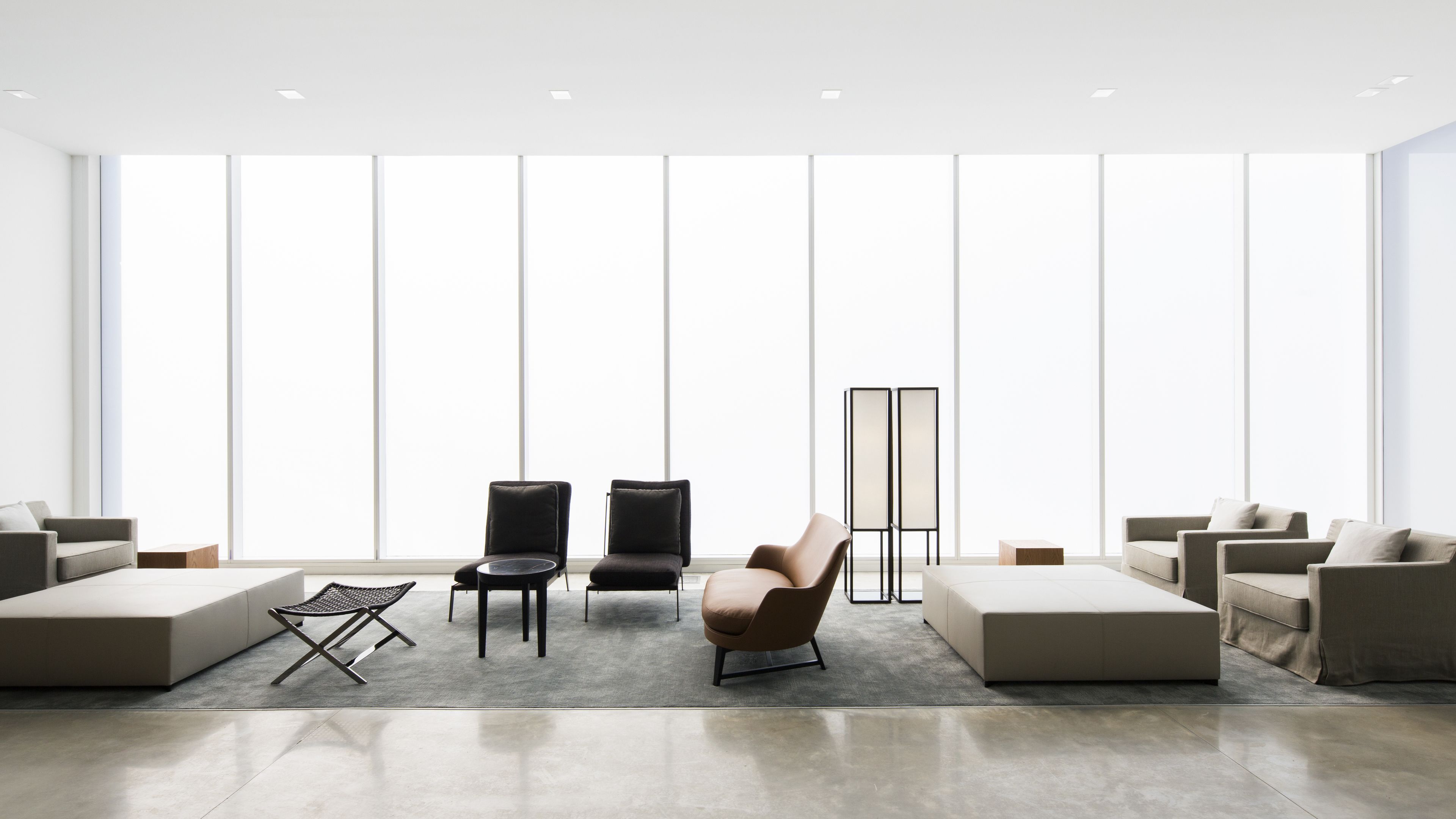 A modern, minimalist living room with large windows, two black chairs, a brown armchair, and a low coffee table.