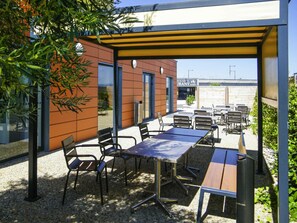An outdoor patio with tables and chairs, a building with a brown facade, and a clear sky. - Ibis Budget Castelnaudary (Castelnaudary)
