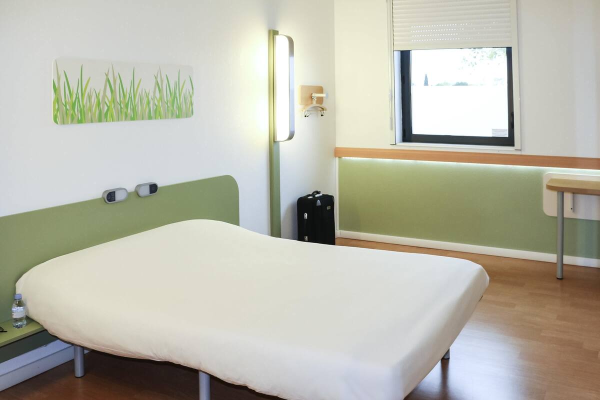 Quarto de casal com cama de solteiro, cabeceira verde, mesa de cabeceira com um frasco, luminária na parede e janela com persianas.