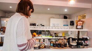 Una persona preparando comida en una cocina con una cafetera, frascos de condimentos y un estante con vasos.