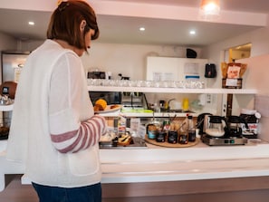 Uma pessoa preparando comida em uma cozinha com uma máquina de café, potes de condimentos e uma prateleira com copos.