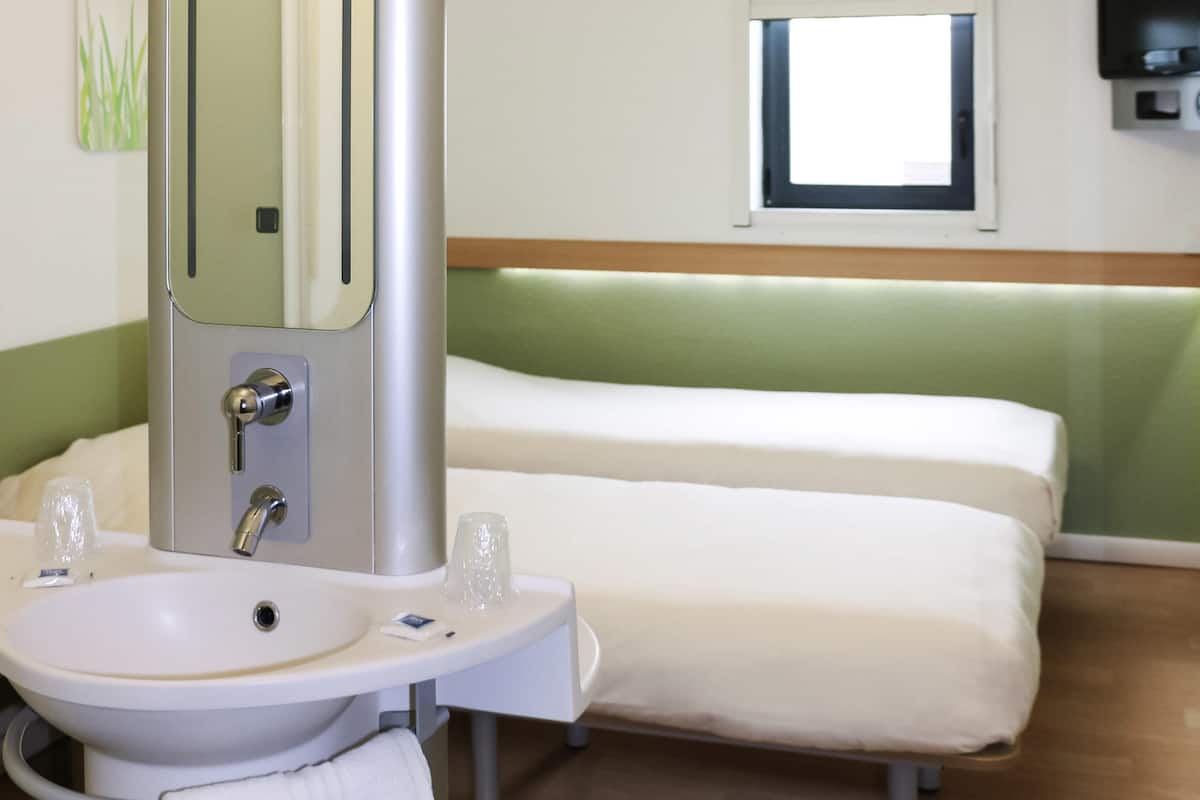 Quarto de hotel com cama, mesa de cabeceira com espelho, pia e janela com persianas.