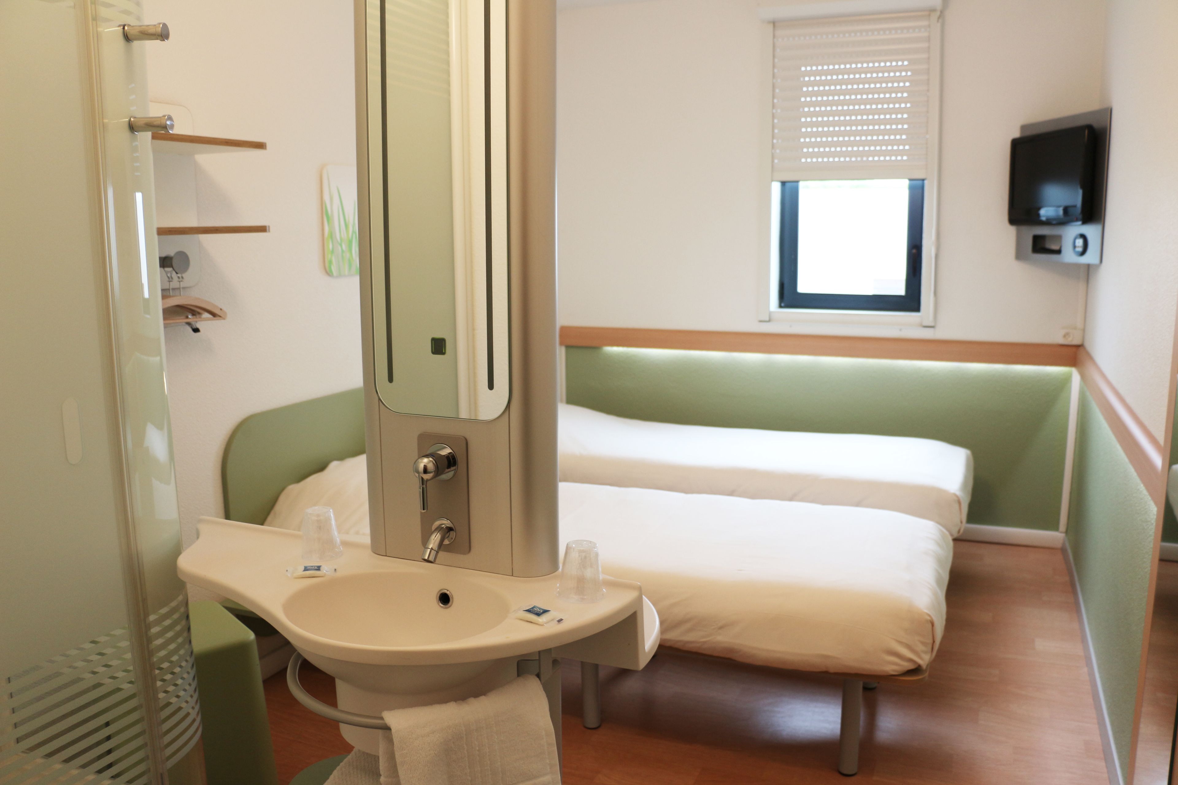 A hotel room with a bed, a bedside table with a mirror, a sink, and a window with blinds.