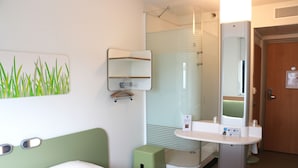 Habitación de hotel con cama, un cubo de basura verde pequeño, un lavamanos blanco con espejo y zona de ducha.