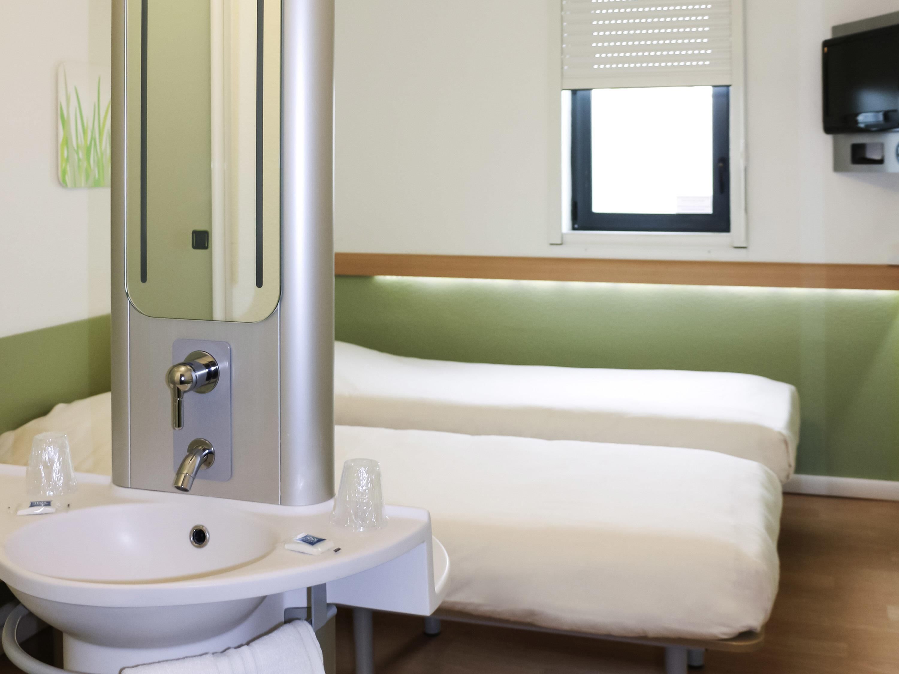 A hotel room with a bed, a bedside table with a mirror, a sink, and a window with blinds.