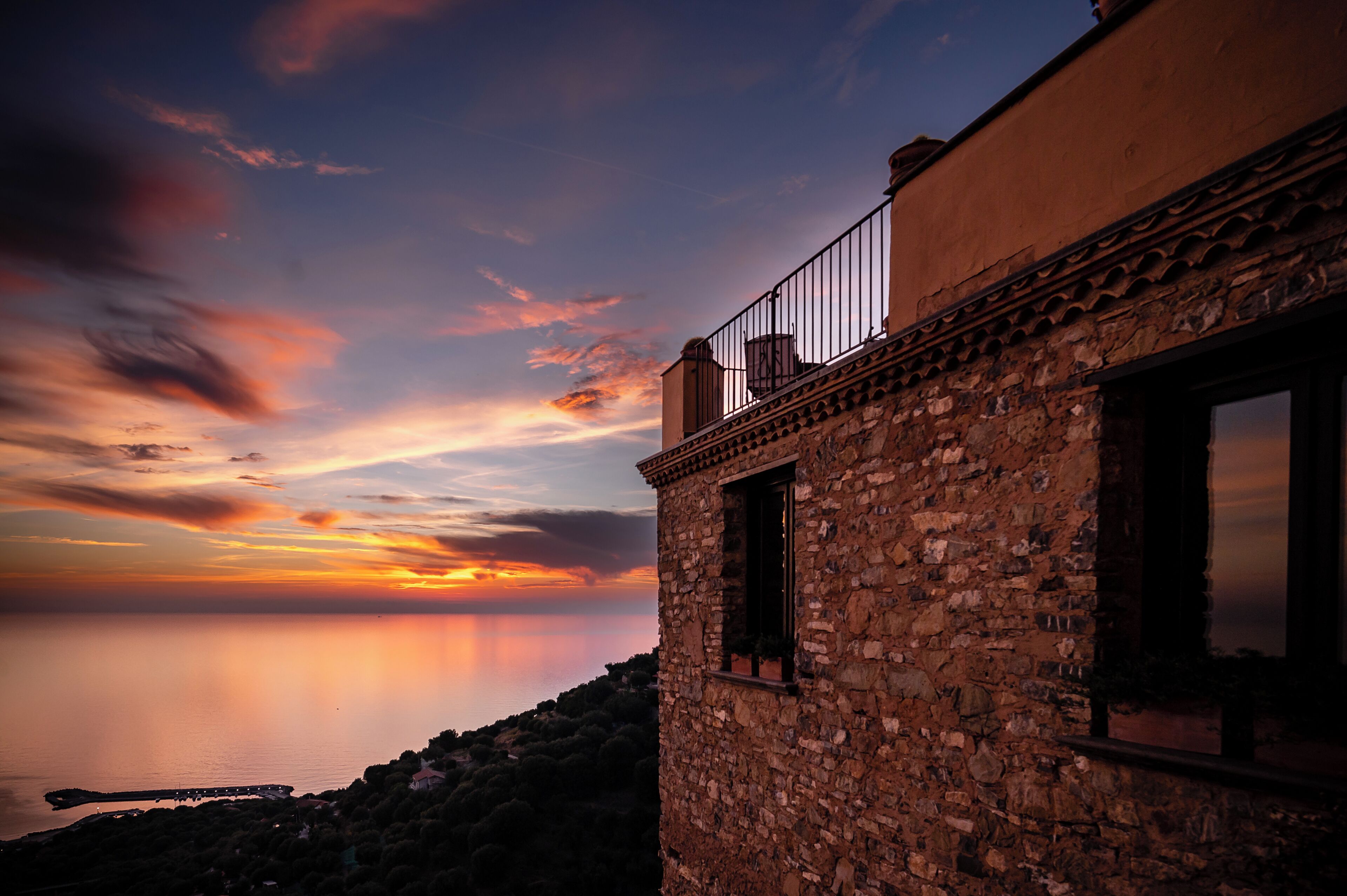 Un bâtiment en pierre doté d’un balcon donnant sur une étendue d’eau au coucher du soleil.