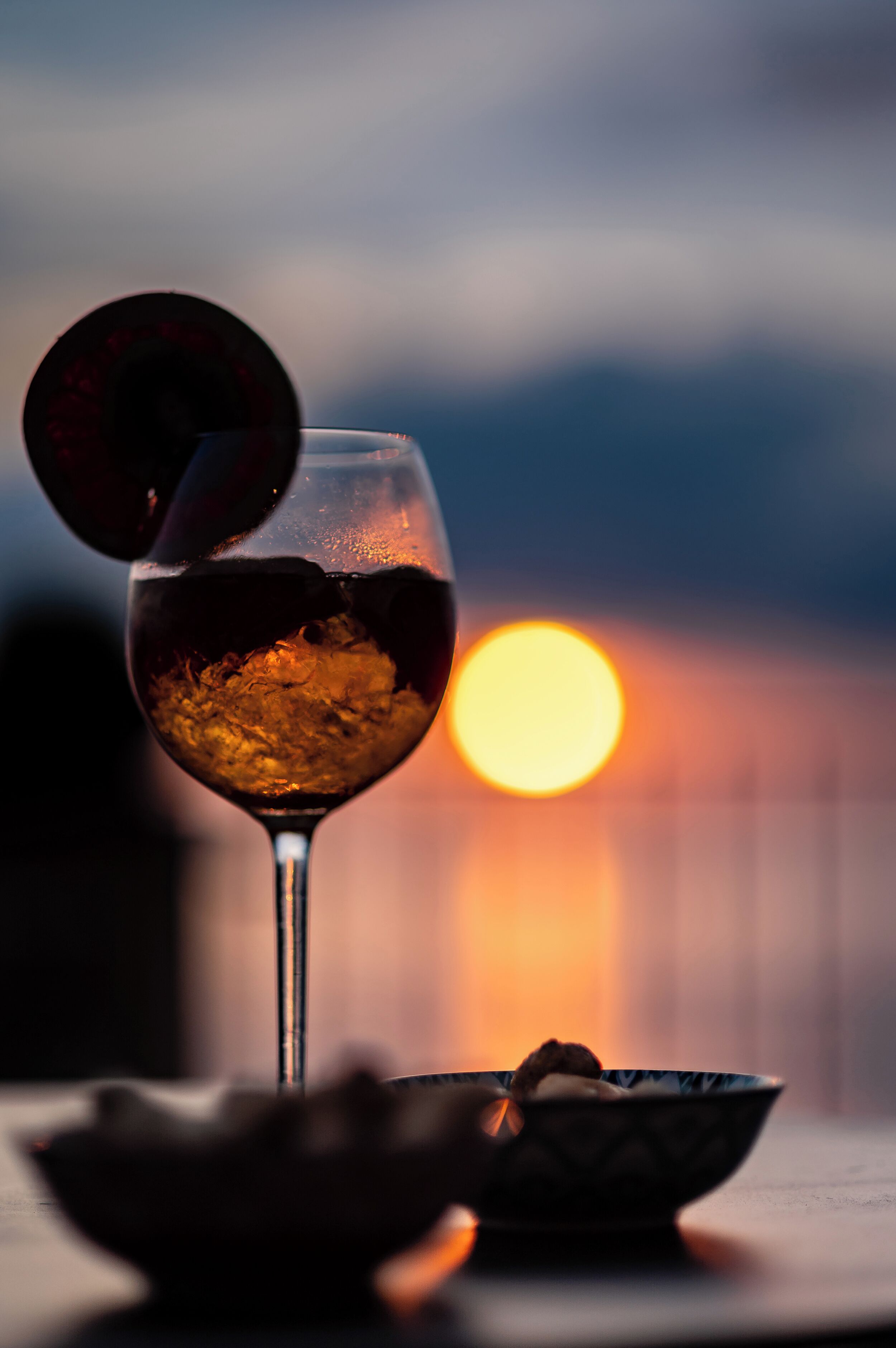 A glass of red wine with ice cubes and a decorative garnish, placed on a table with a bowl in the foreground, against a sunset backdrop.