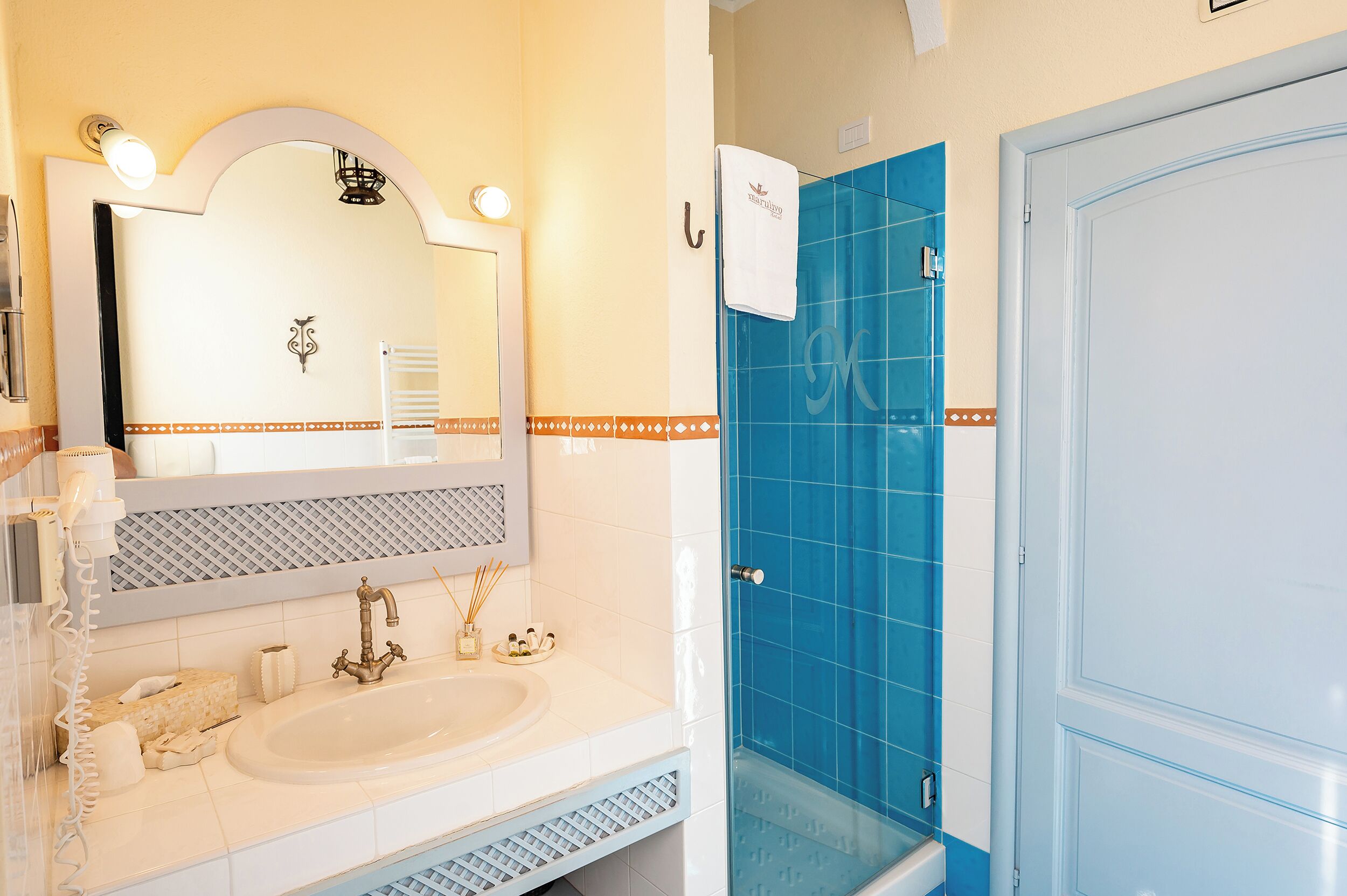 A bathroom with a white sink, a mirror with a decorative frame, a blue shower enclosure, and a white door.