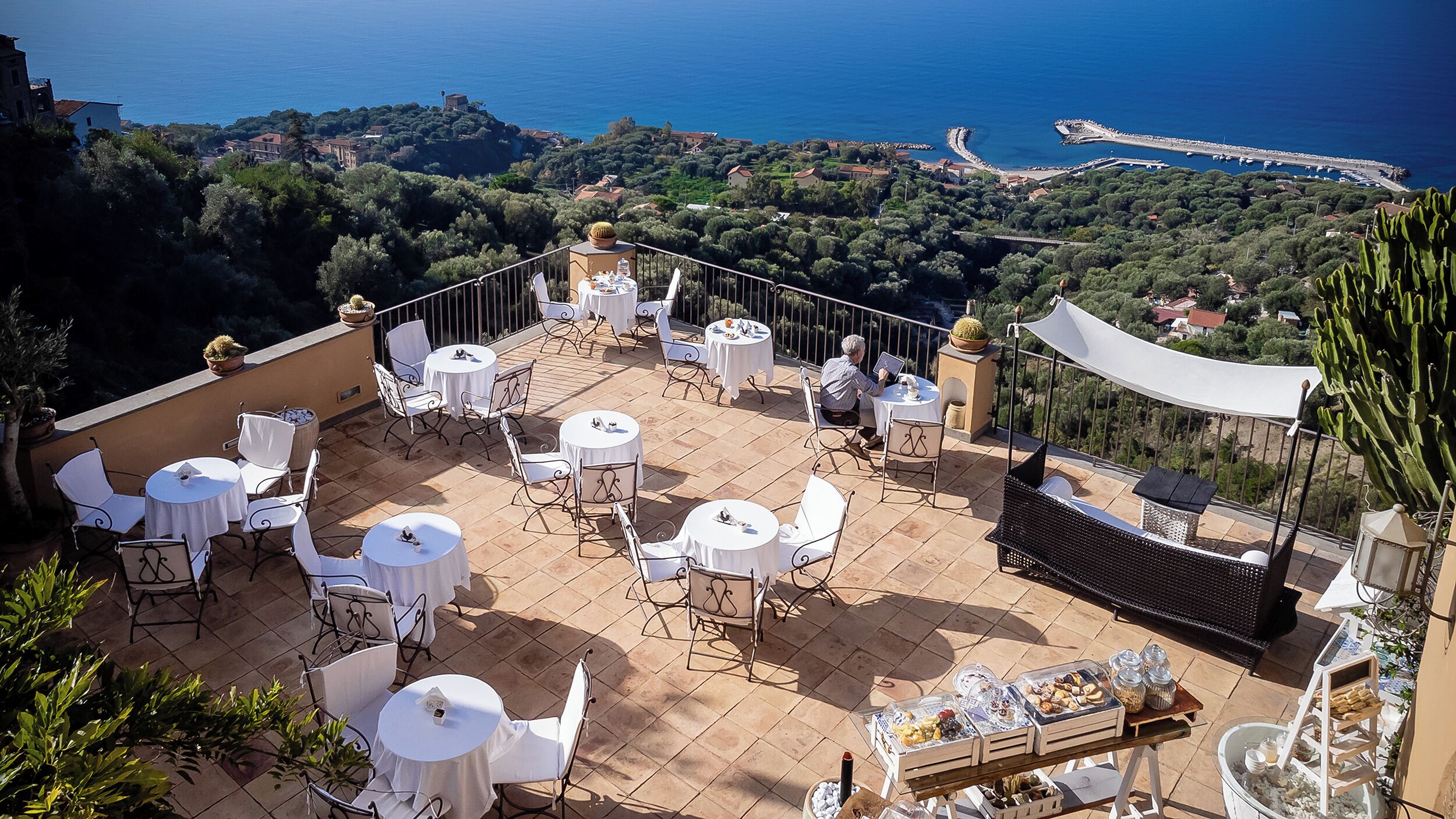 A terrace with round tables and chairs, a cactus, and a view of the sea and hills.