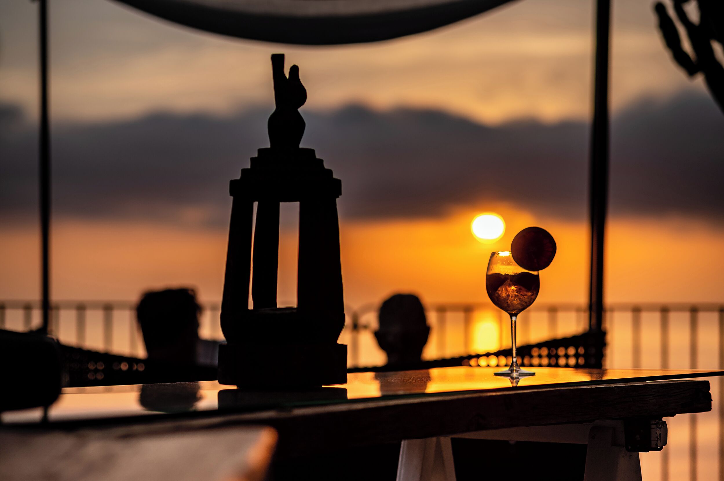 A sunset view from a balcony with a table, a glass of drink, and a statue.