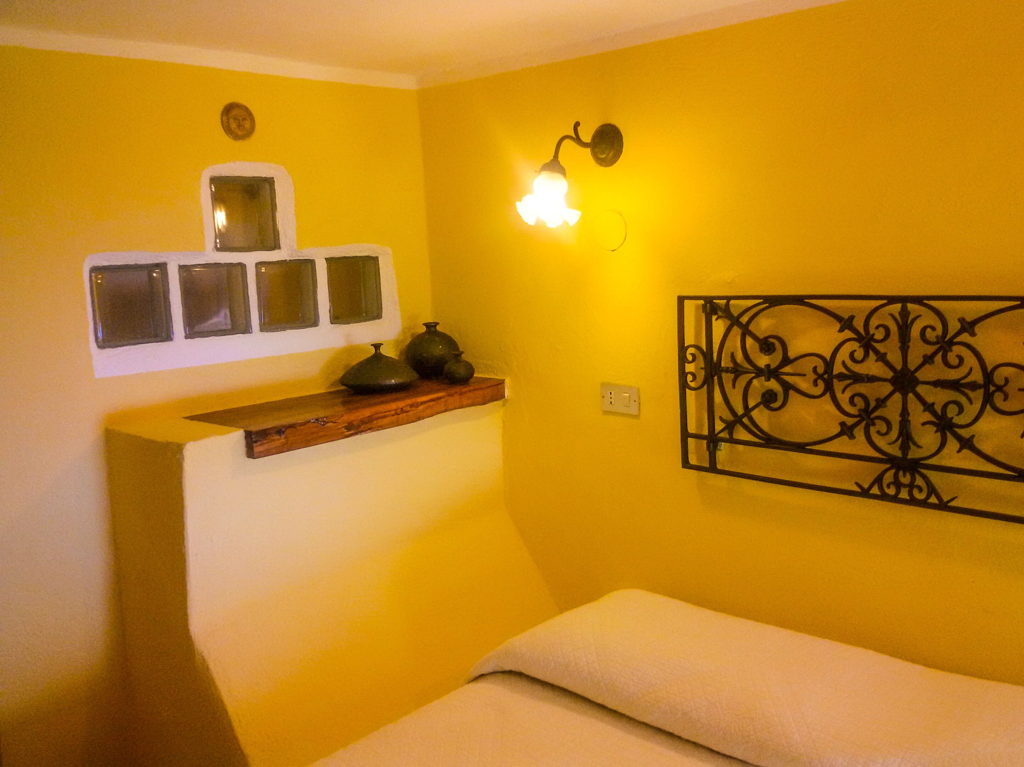 A room with a yellow wall, a wooden shelf with decorative items, a wall-mounted light fixture, and a metal wall art piece.