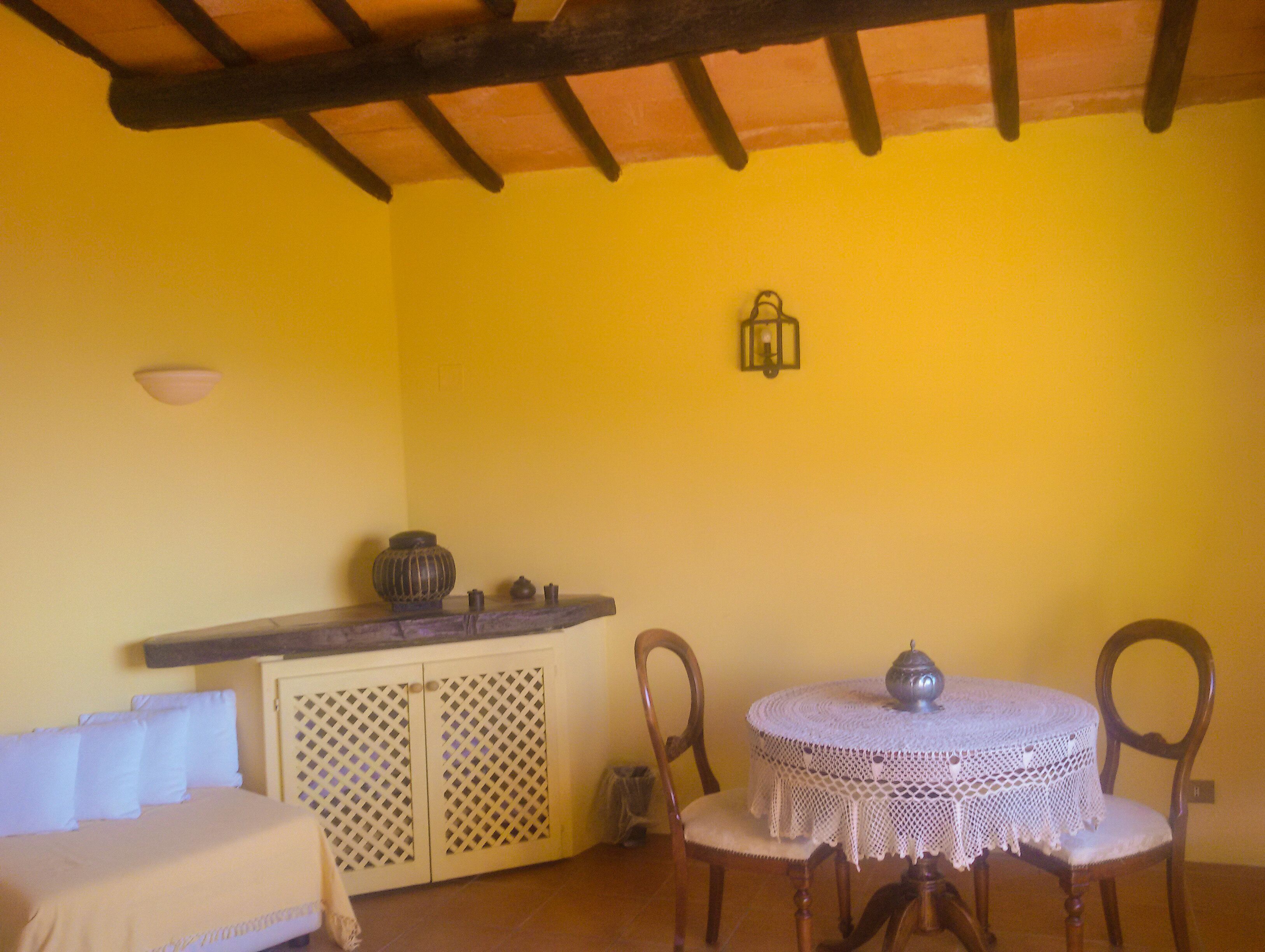 Una stanza con una parete gialla, un soffitto in legno, un letto con biancheria bianca, un tavolino con tovaglia all'uncinetto e una sedia.