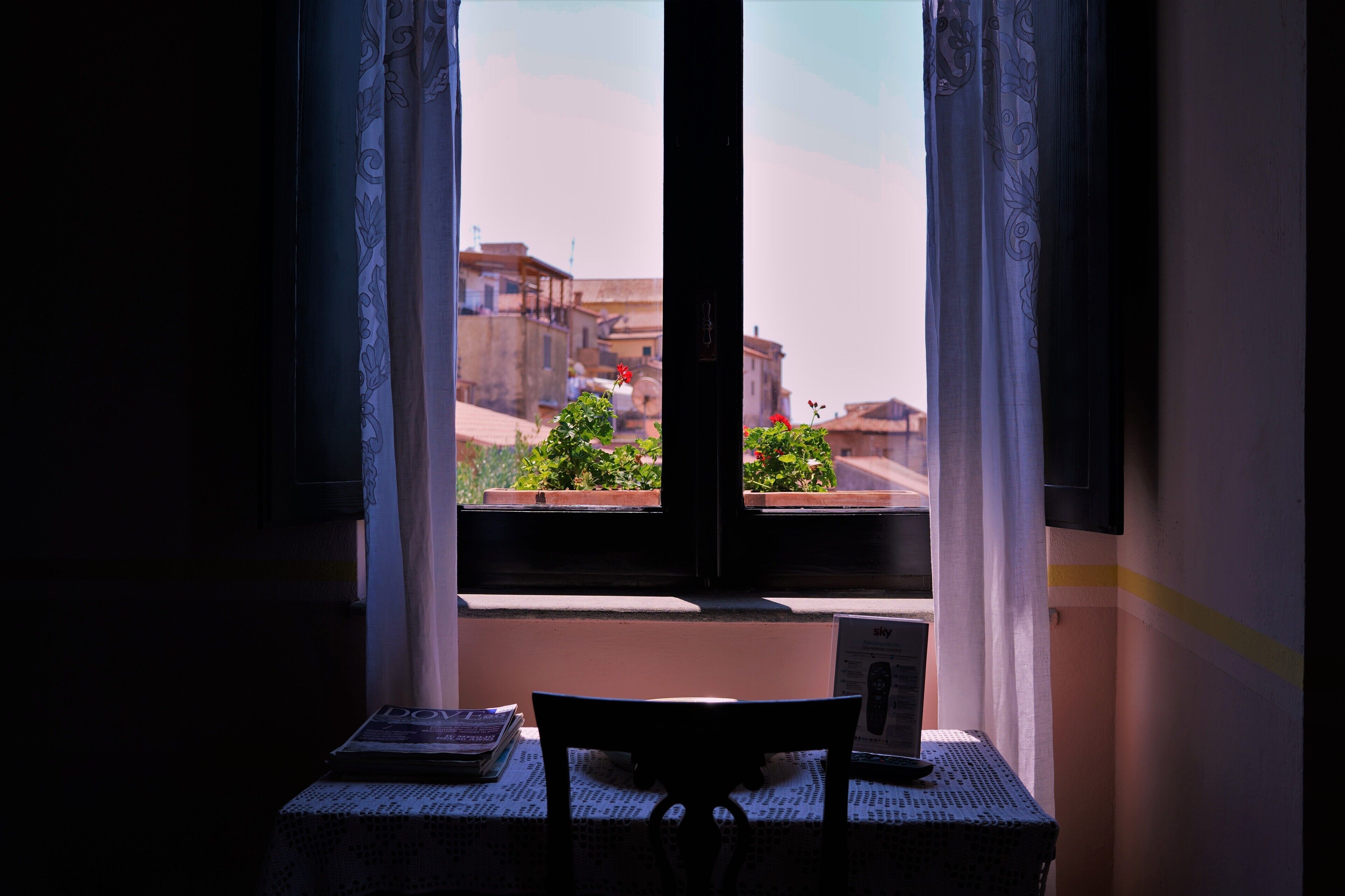 Une pièce avec une fenêtre donnant sur un paysage urbain, une table avec une chaise, et un petit bureau avec une lampe et un livre.