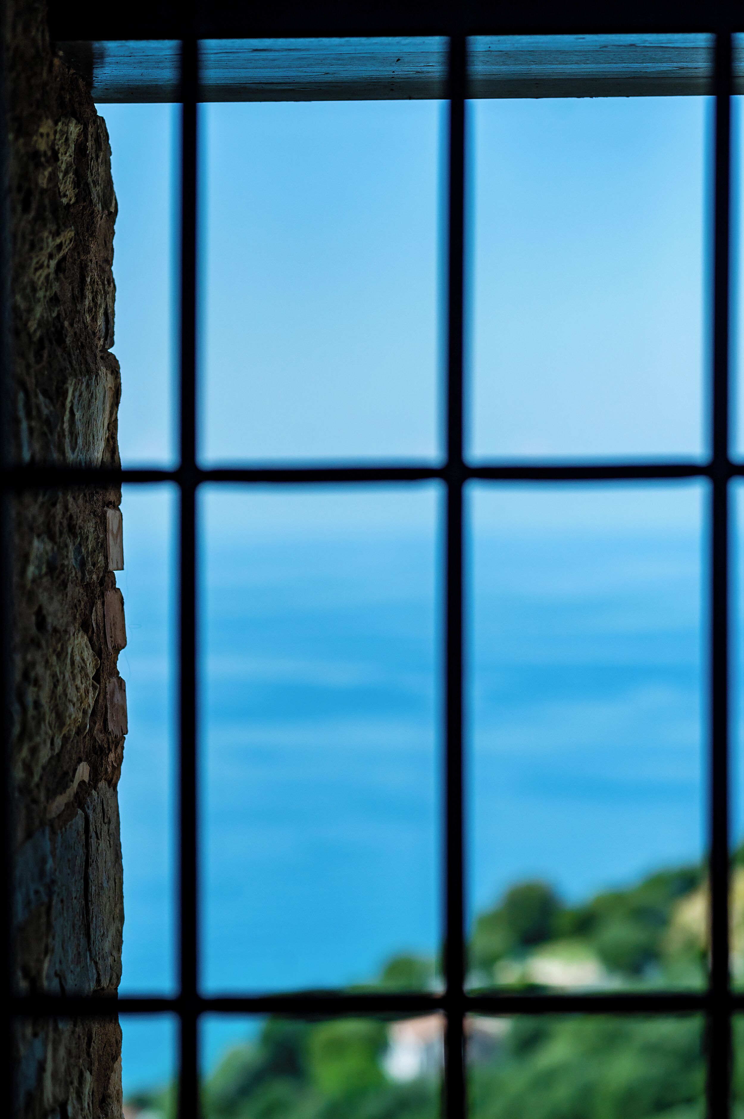 A window with a stone wall on one side and a view of the sea through a grid pattern.