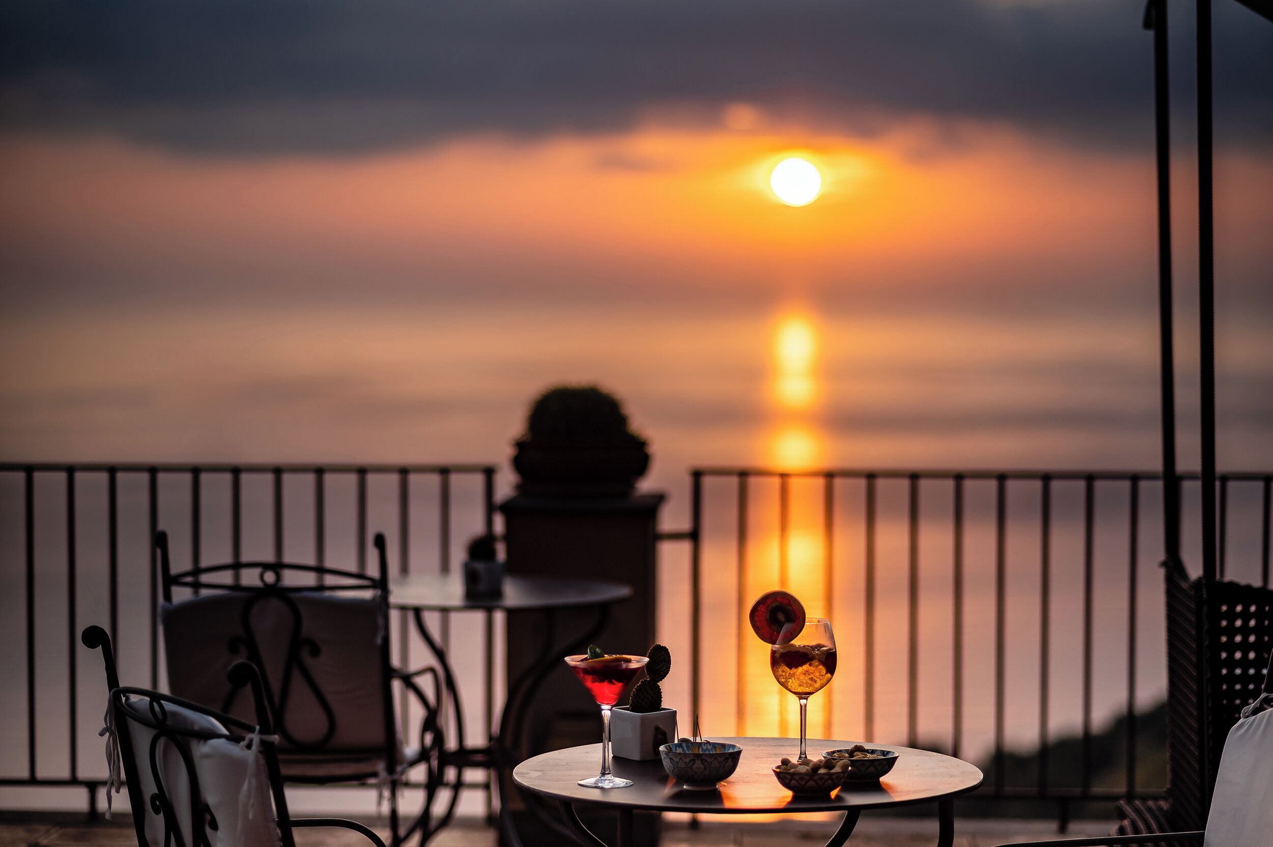 A sunset view from a balcony with a table set for two, featuring drinks and a bowl of fruit.